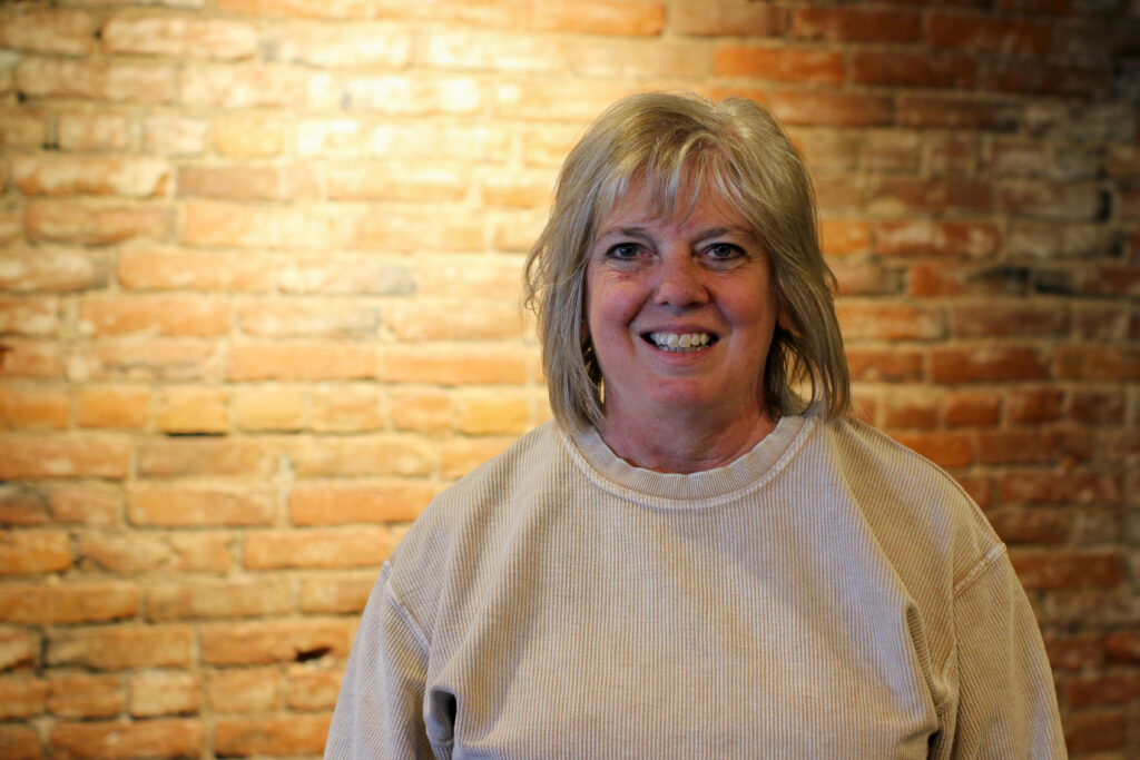 A woman poses in front of a brick wall brightened by an overhead light and smiles at the camera. She has shoulder length blonde hair and is wearing a cream colored sweater.