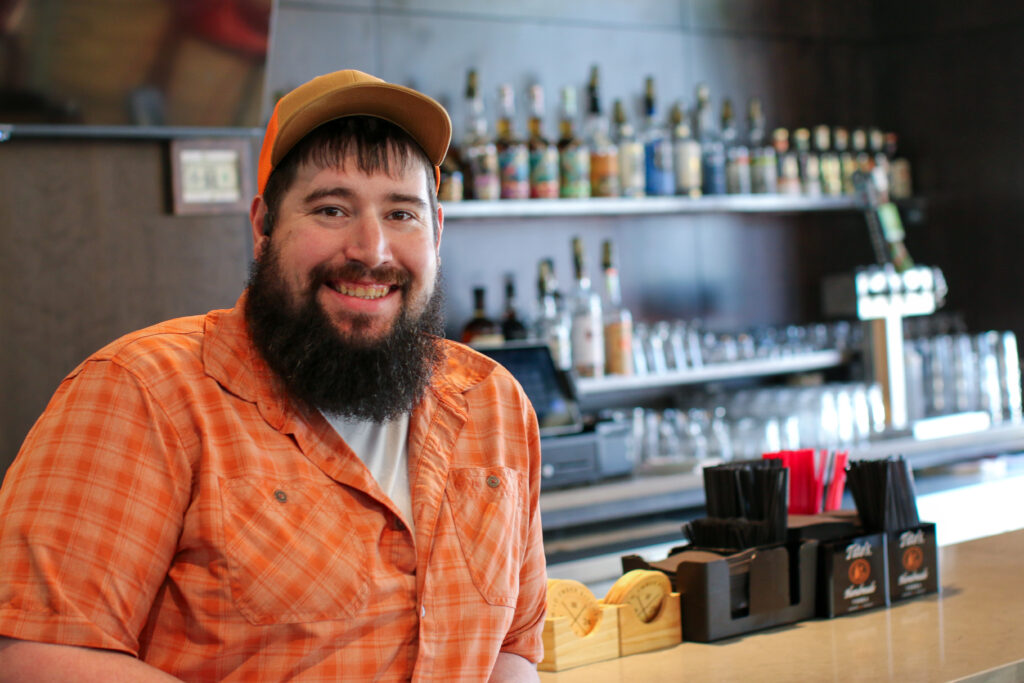 A man leans against a bar top with liquor bottles, beer tap and glassware visible behind him. He's wearing a green shirt and ball cap, has a full beard, and is smiling at the camera.