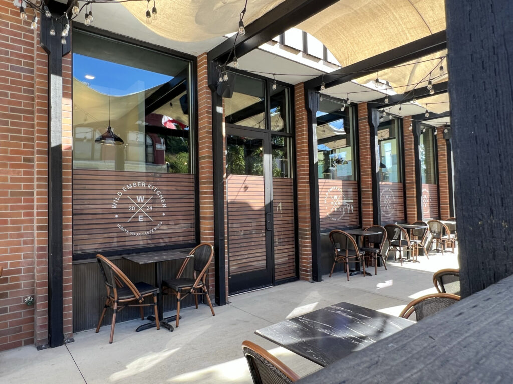 An outdoor dining patio with small tables with two chairs lining opposite walls. A canopy and string lights are visible above. Dark beams and the brick walls of the building frame large windows with the Wild Ember logo.