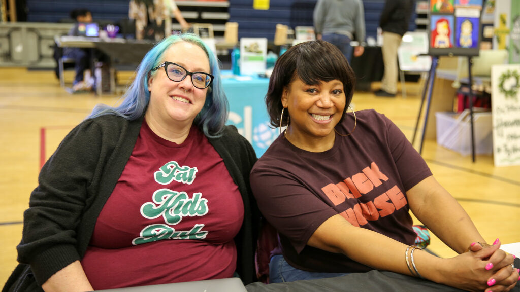 A white woman and Black woman pose together at a table in a gymnasium. Other tables and displays are visible in the background. 