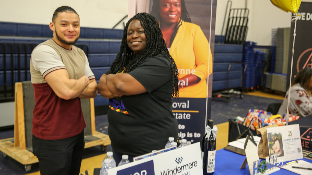 A man and woman pose together with their arms crossed in a gymnasium. A pop-up sign with an image of the woman is displayed behind them and a table is visible in the foreground.