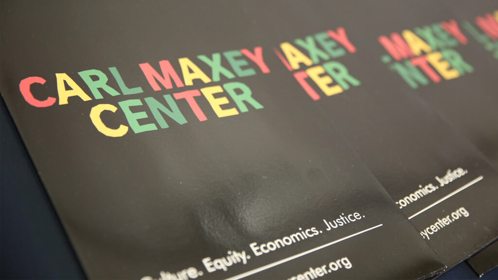 Black folders with "Carl Maxey Center" in red, yellow and green font lay in a line on a dark background. "Culture. Equity. Economics. Justice." is visible in white on the bottom of the folders.