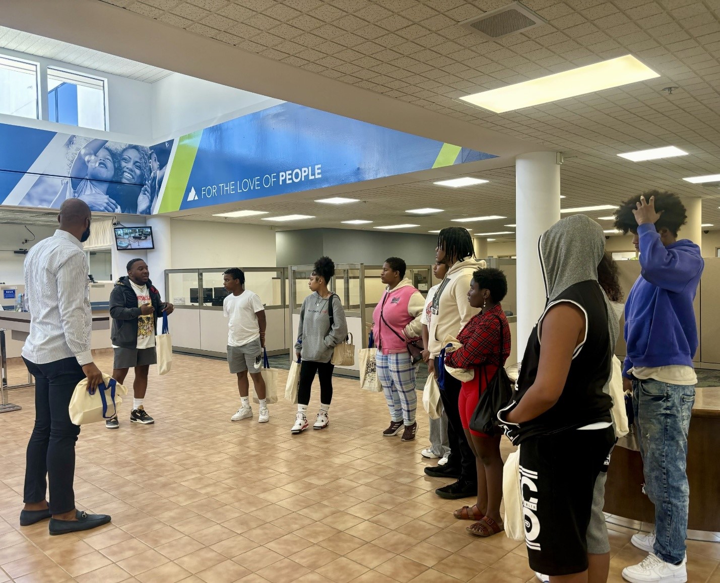 A group of young people listen to a man speak in the lobby of a credit union in Tacoma. One man is wearing business casual attire, while the others are in casual attire, including shorts and hooded sweatshirts. A blue and green banner stretches above them, windows let in natural light, and cubicles and teller stations are visible in the background.