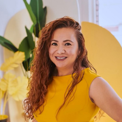 Noemi Cagatin-Porter, founder of CJK Community Homes, poses in natural light. Her long hair is draped over one shoulder as she smiles at the camera. She is wearing a yellow top. Yellow accents and greenery are visible in the background