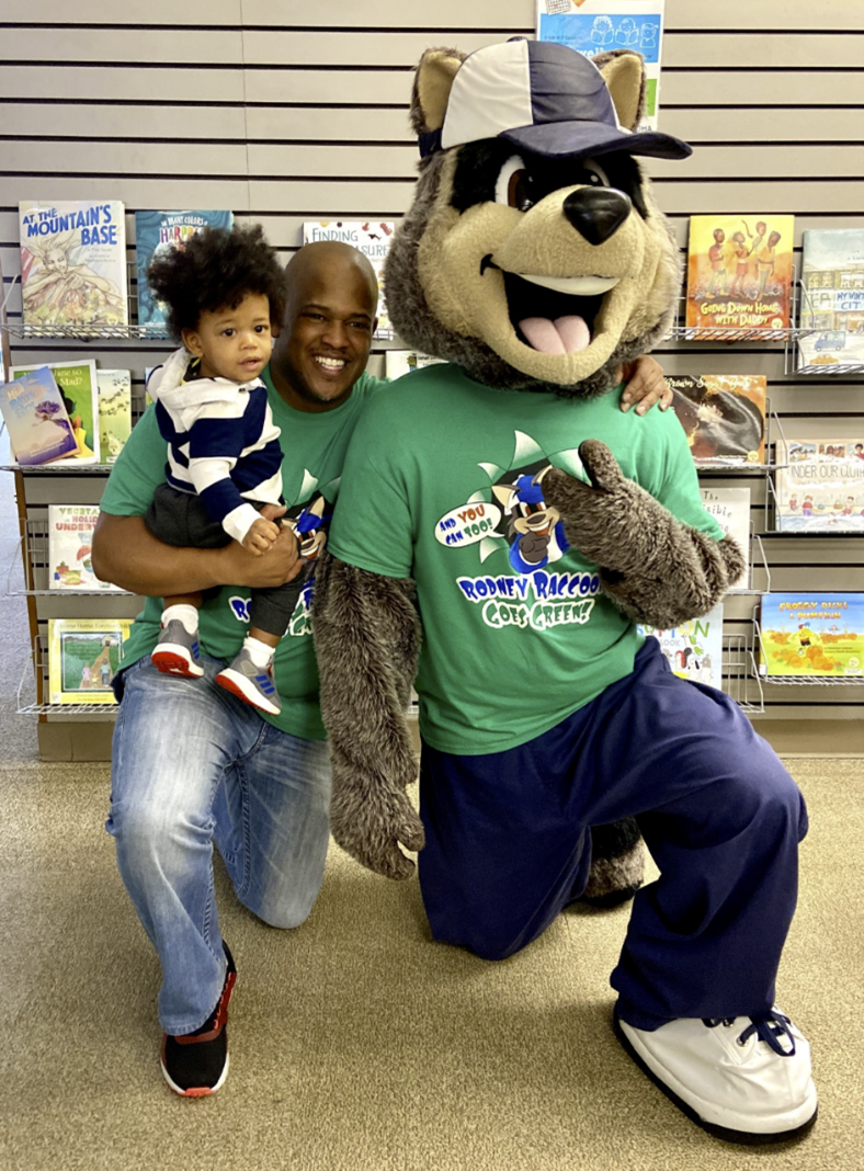 Frederick “JD” Davis and his son pose with Way Out Kids mascot Rodney Raccoon at a children’s book signing at the Tacoma Public Library.